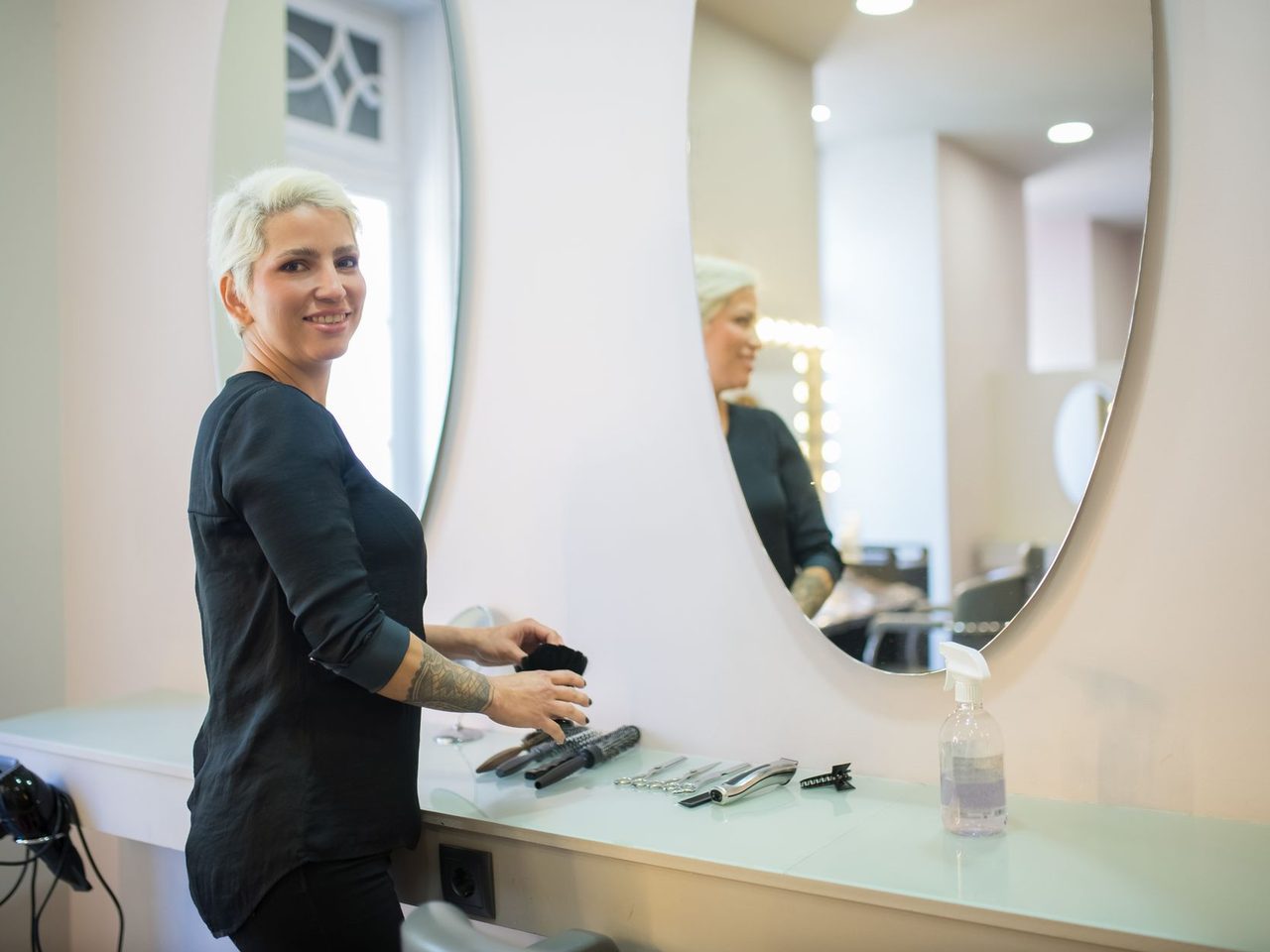 Woman with short blonde hair standing at a white salon counter with hair tools, beside a large round mirror in a bright room.