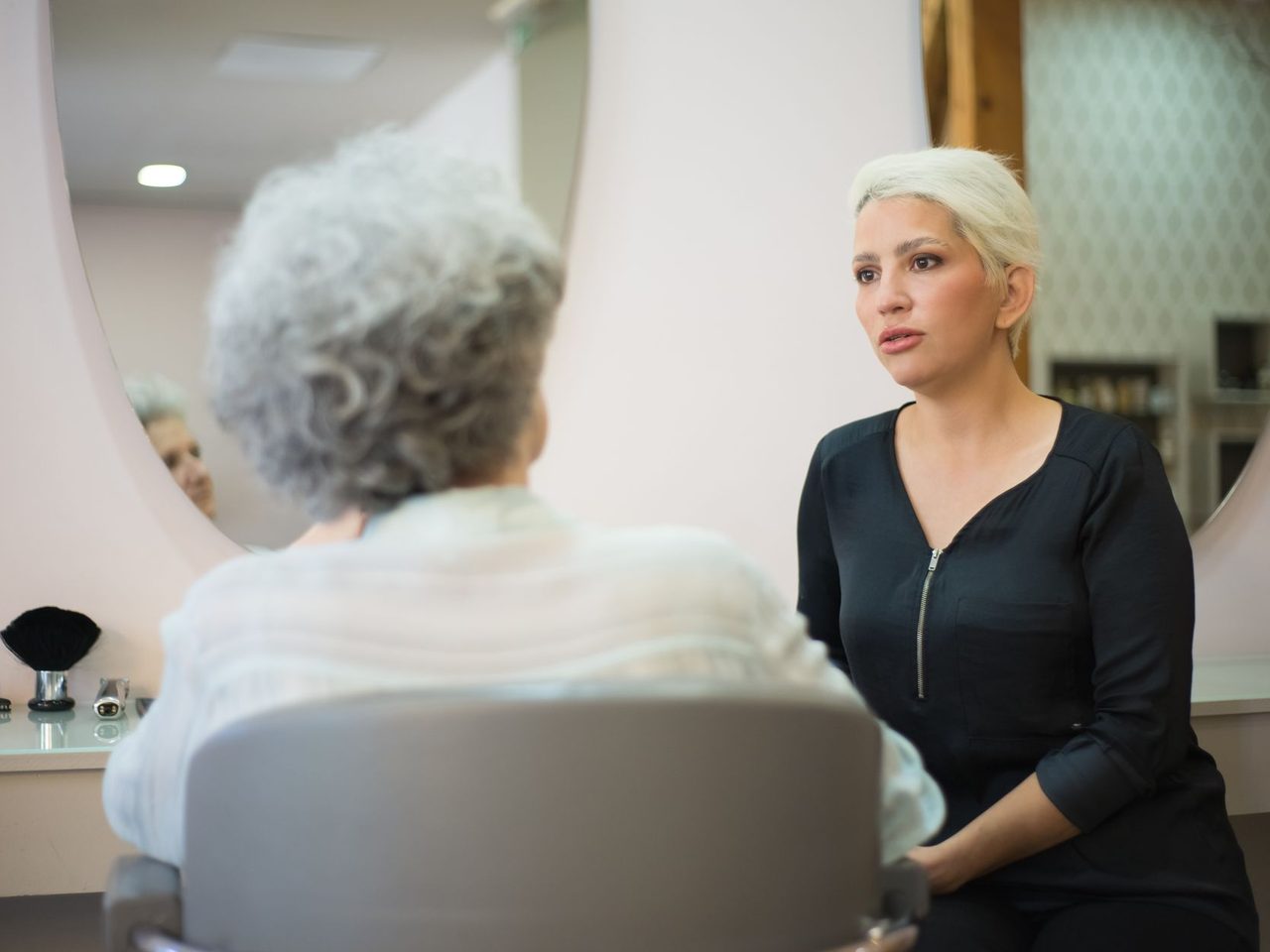 Woman with short gray hair seen from behind sitting in a salon chair while a blonde stylist in black stands beside her talking.