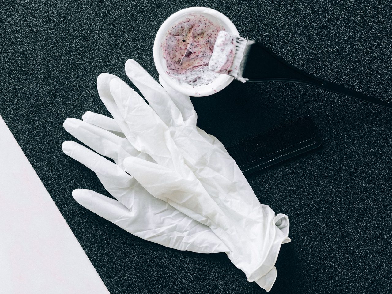 Flat lay of white latex gloves, a black tint brush, a small bowl of pink hair dye and a comb on a dark background.