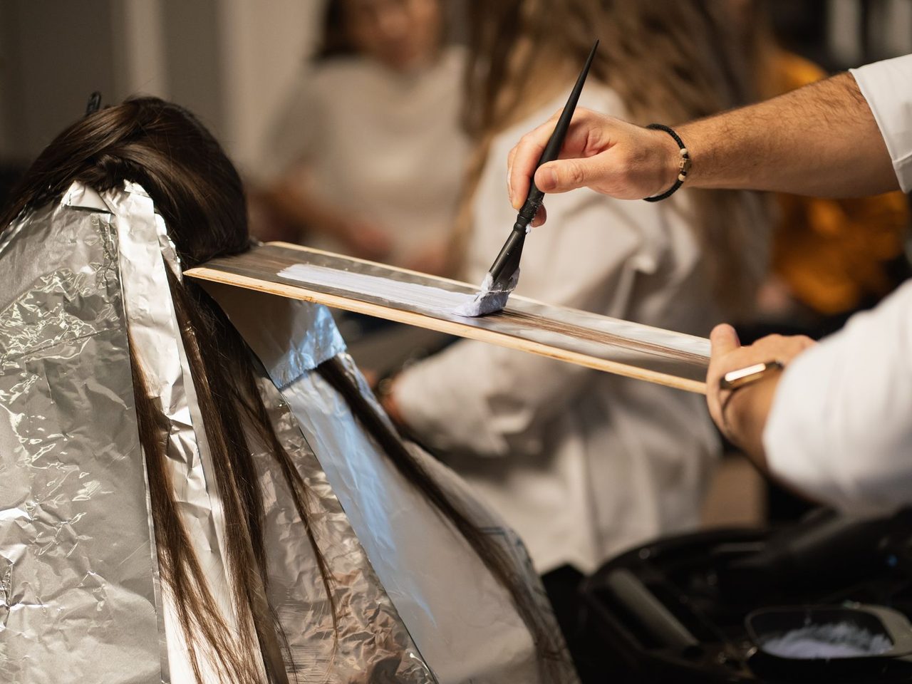 Client seen from behind with long dark hair sectioned over aluminum foil while a hairdresser applies dye with a brush and paddle.