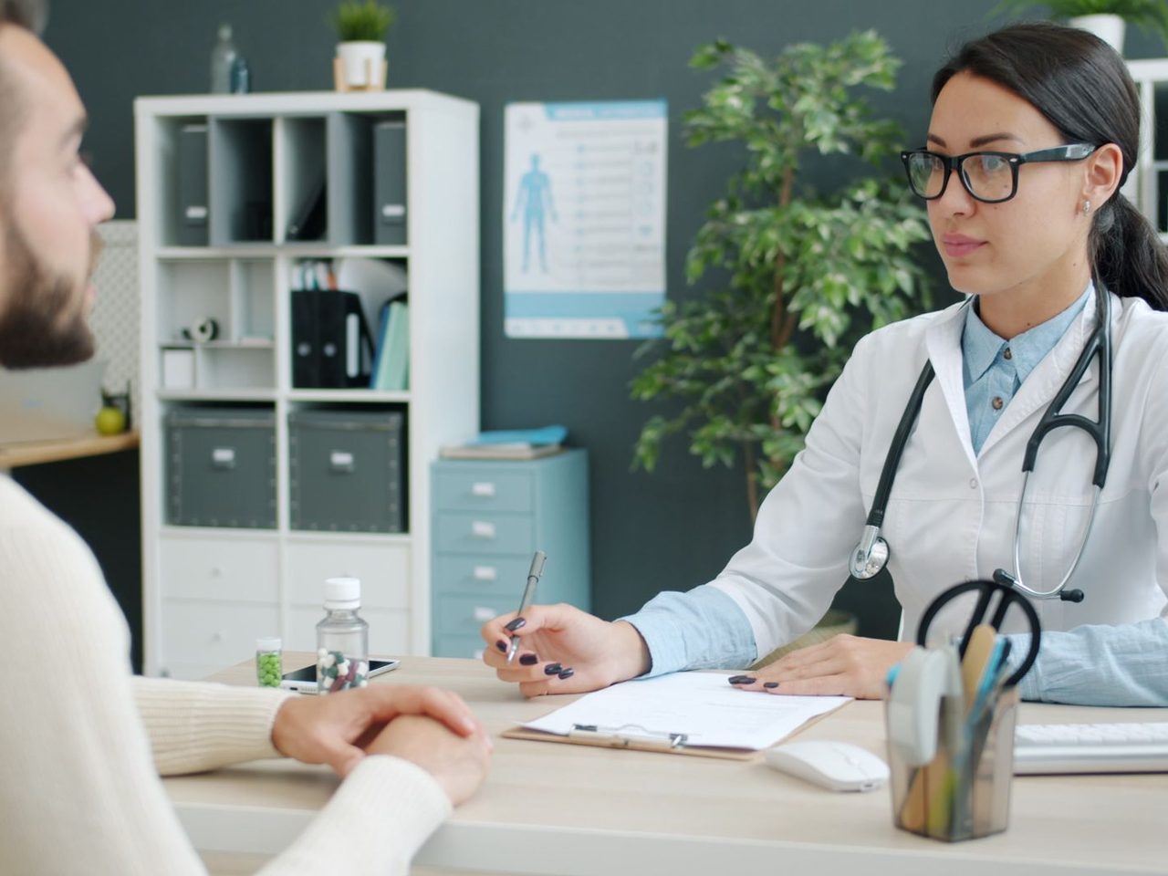 A doctor in a white coat with a stethoscope listening to a male patient across a desk