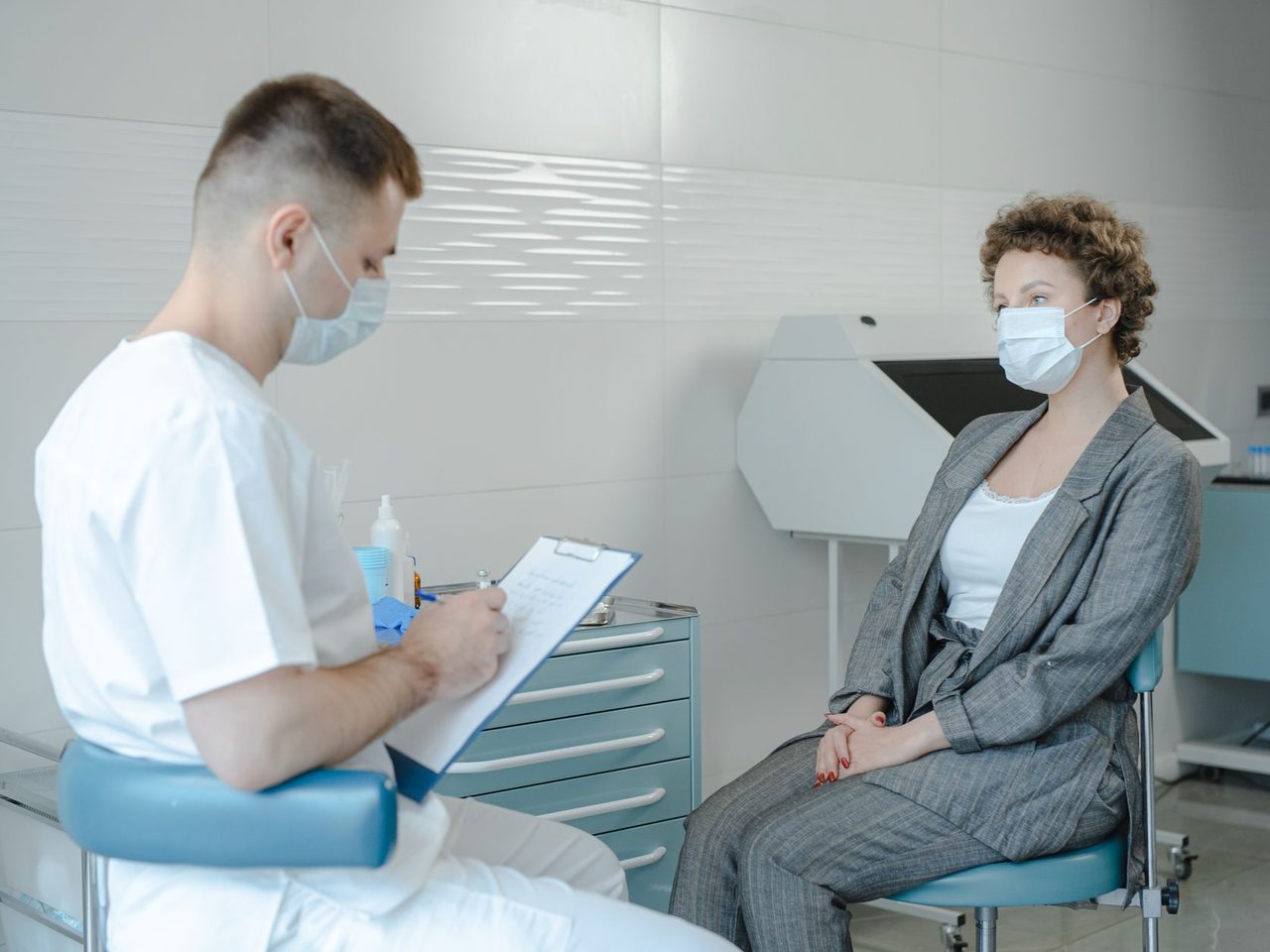 A doctor in a face mask taking notes while talking with a seated woman patient in a clinic