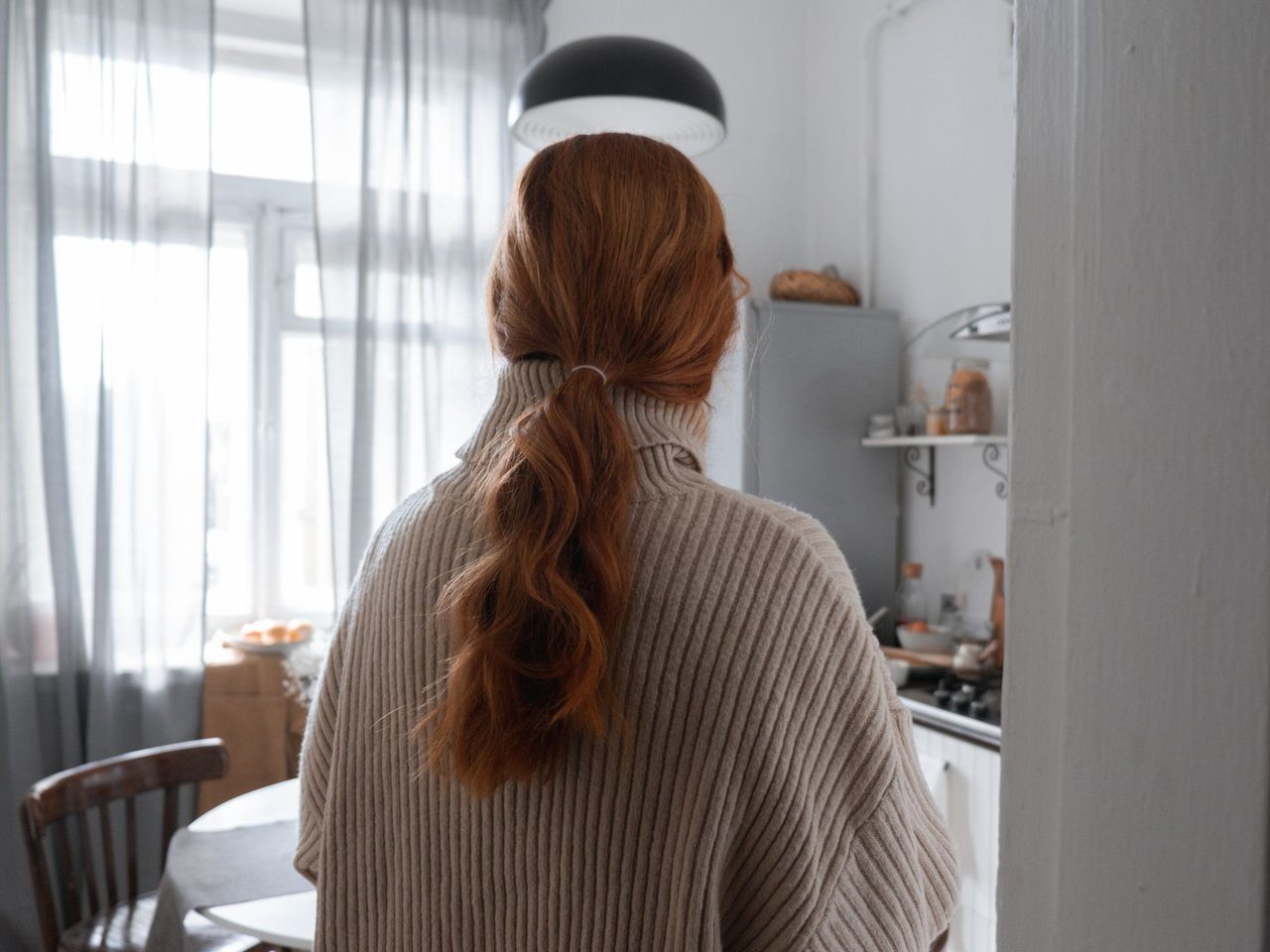 A woman with red hair in a ponytail, seen from behind in a bright kitchen