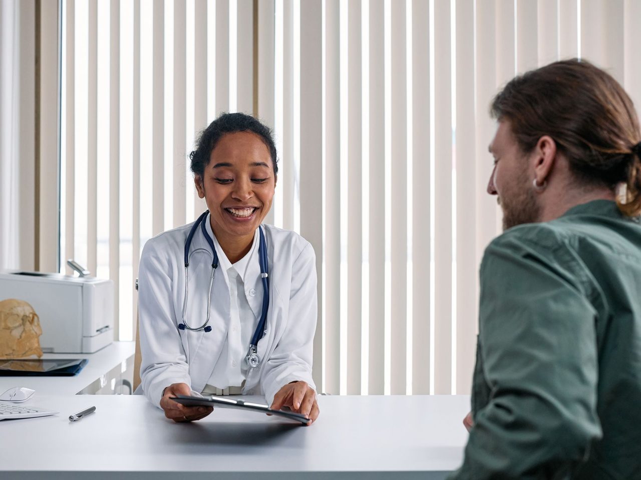 A smiling doctor talking with a male patient across a desk in a bright clinic