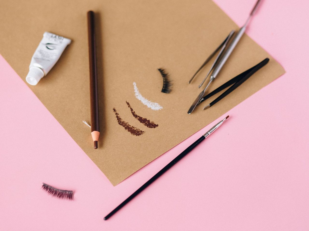 Flat lay of eyebrow pencils, tweezers, a brush, a small white tube and drawn brow strokes on brown paper on a pink background