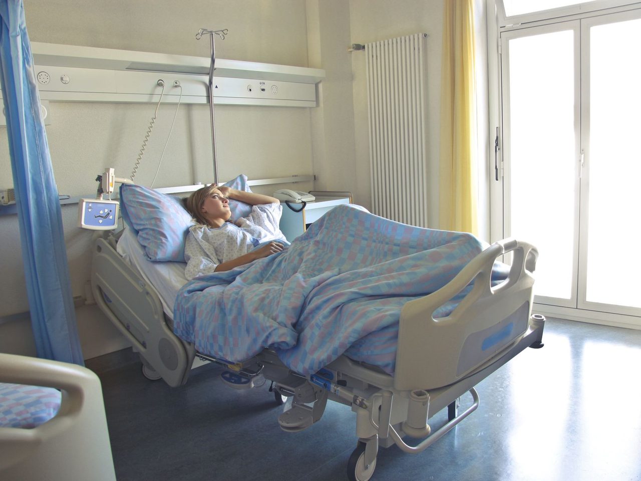 A woman resting in a hospital bed in a bright room with an IV stand beside her