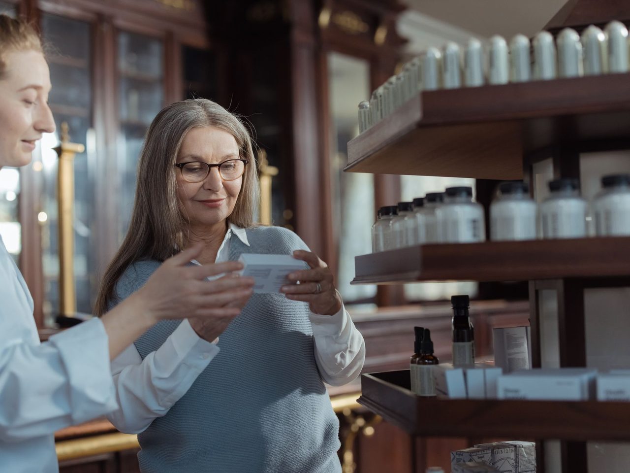 An older woman with glasses talking with a pharmacist among shelves of products