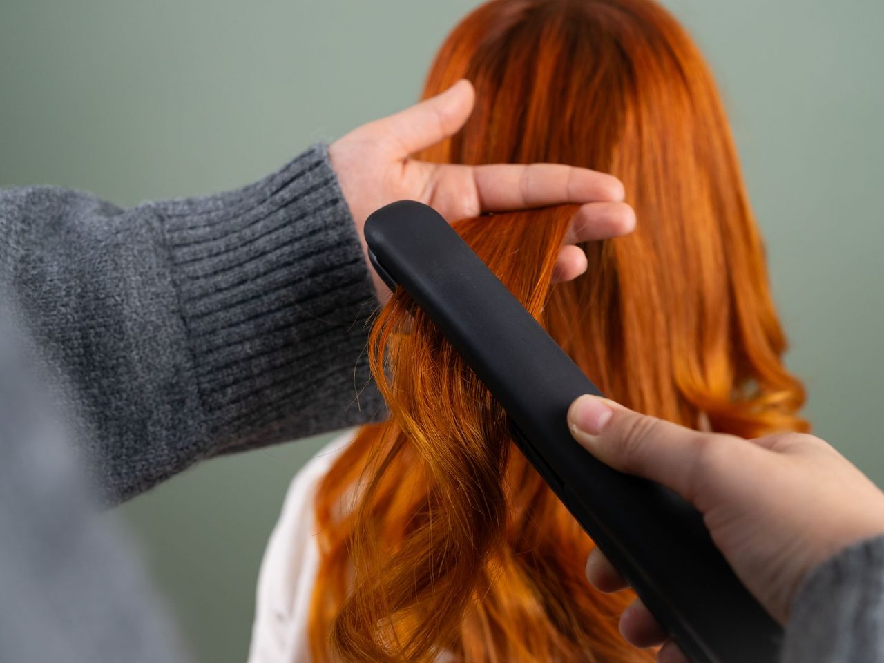 Hands in a gray sweater pressing a black flat iron along a section of long straight red hair against a green background.