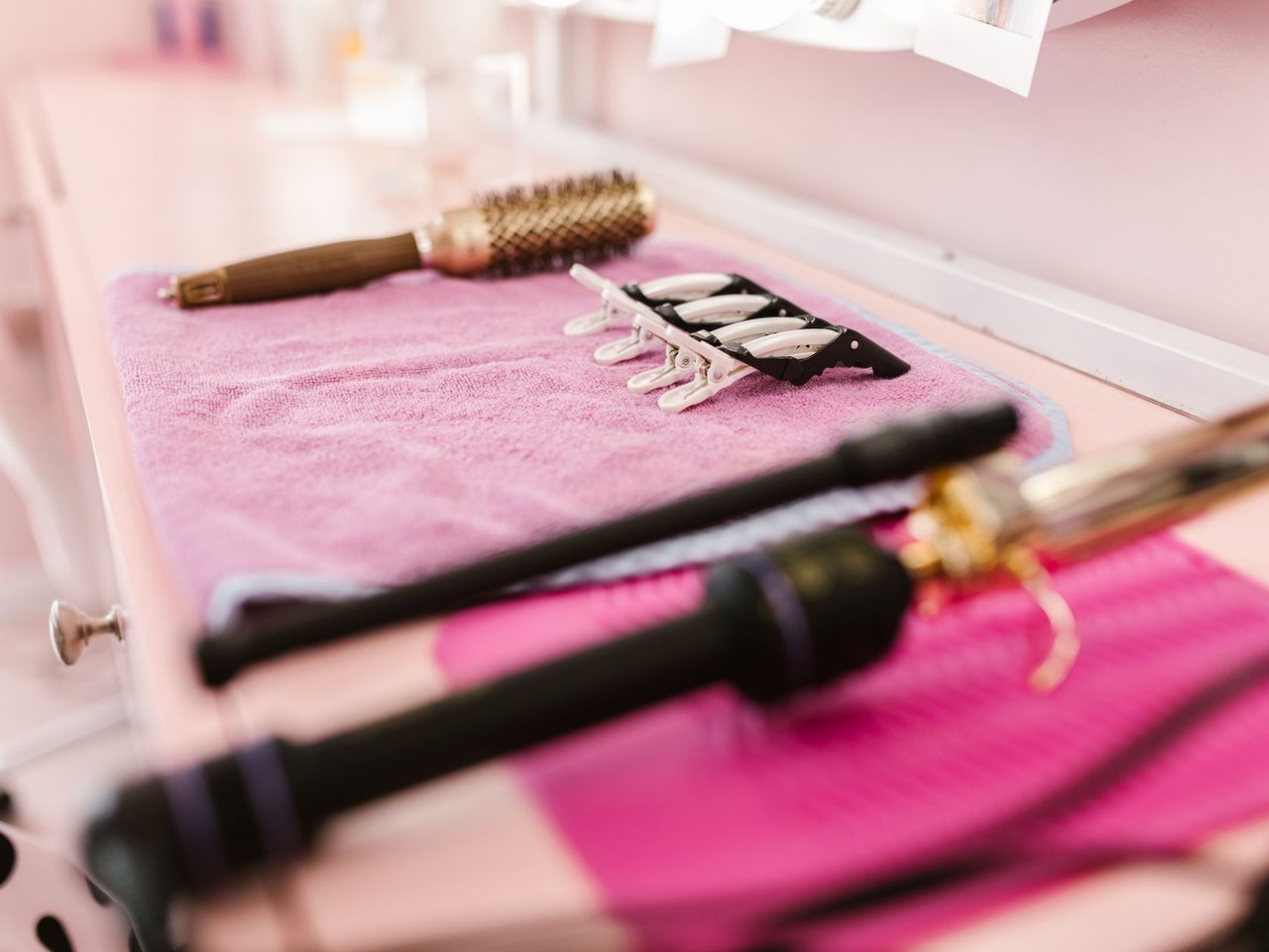 Several heated hair styling tools, including curling irons and a straightener, along with brushes arranged on a pink towel on a salon table.
