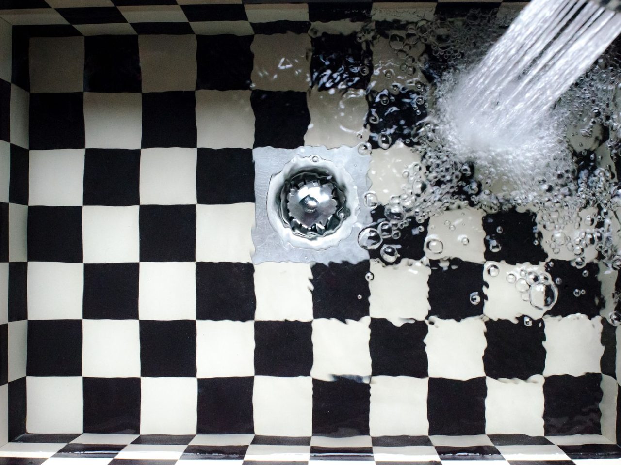 Overhead view of water spraying toward a round drain on black and white checkered tiles
