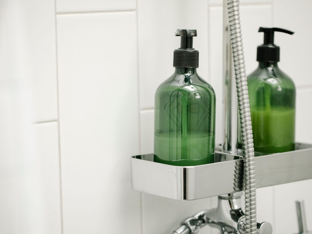 Two green pump bottles on a metal shower shelf against white tiles