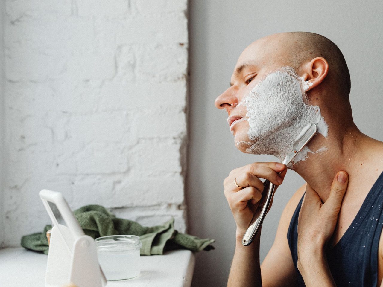 A bald man applying shaving cream to his face and shaving at a bathroom mirror