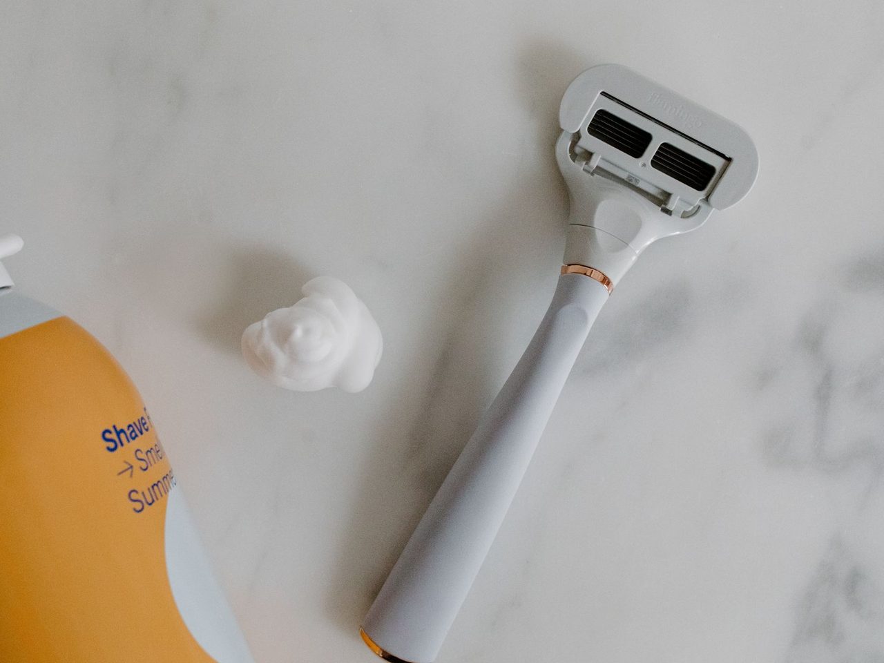 A razor and a can of shaving foam with a dab of foam on a white marble surface