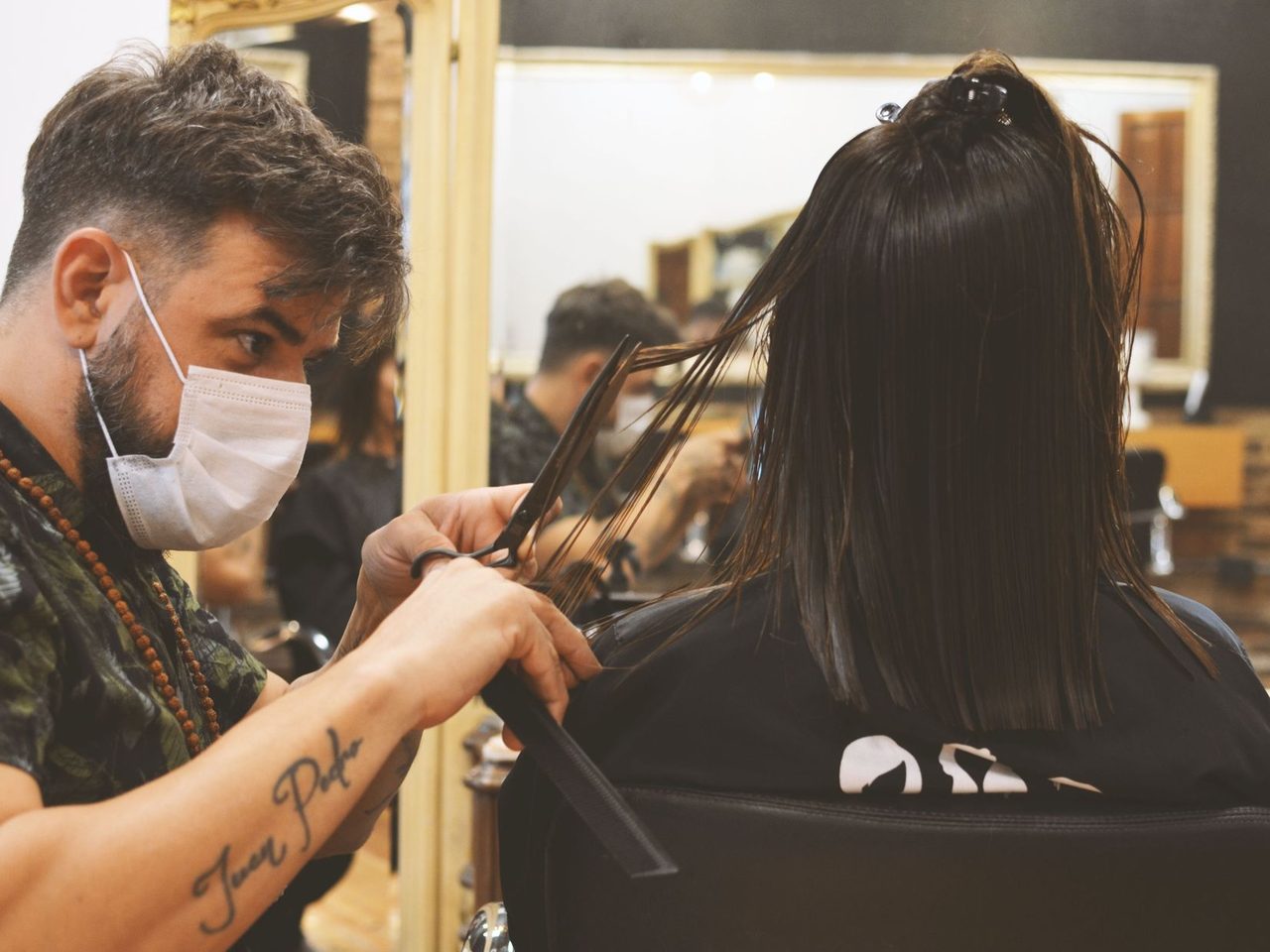 A stylist in a face mask cutting a woman's straight dark hair in a salon
