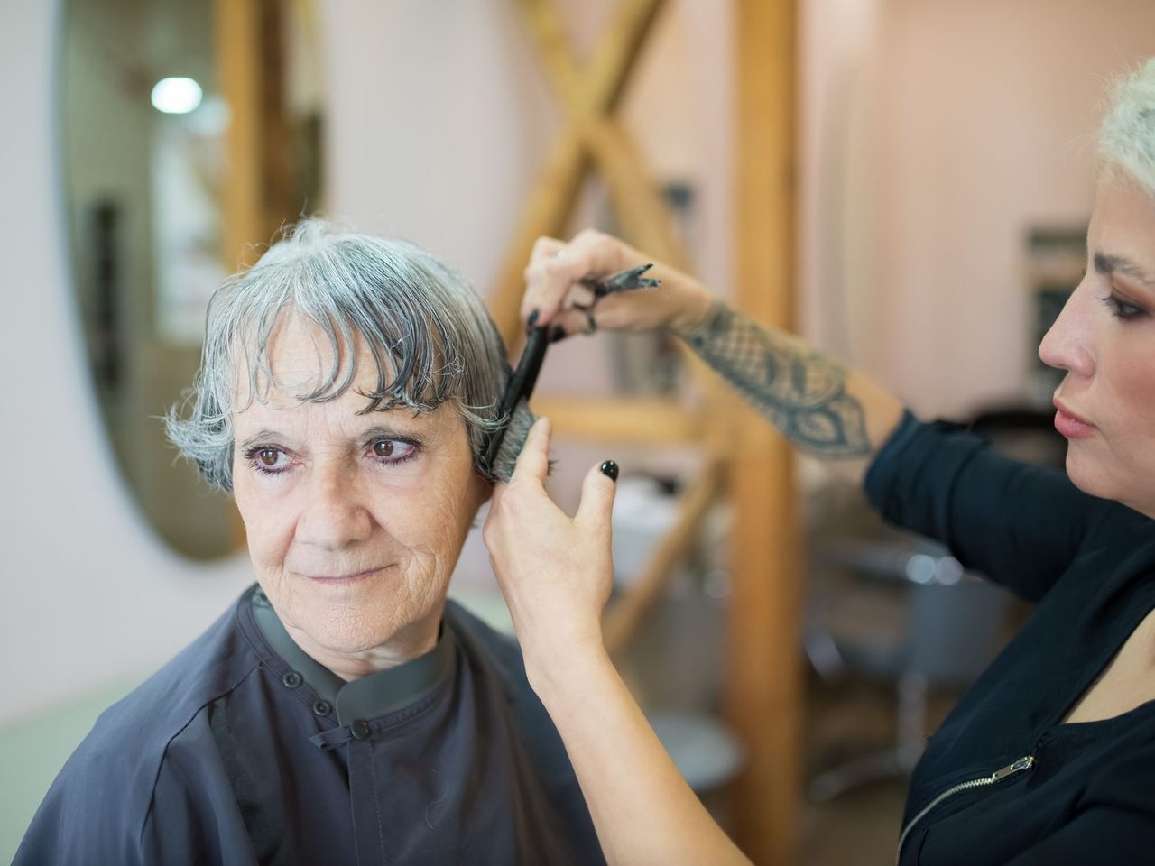 An older woman with short gray hair smiling while a hairdresser styles it