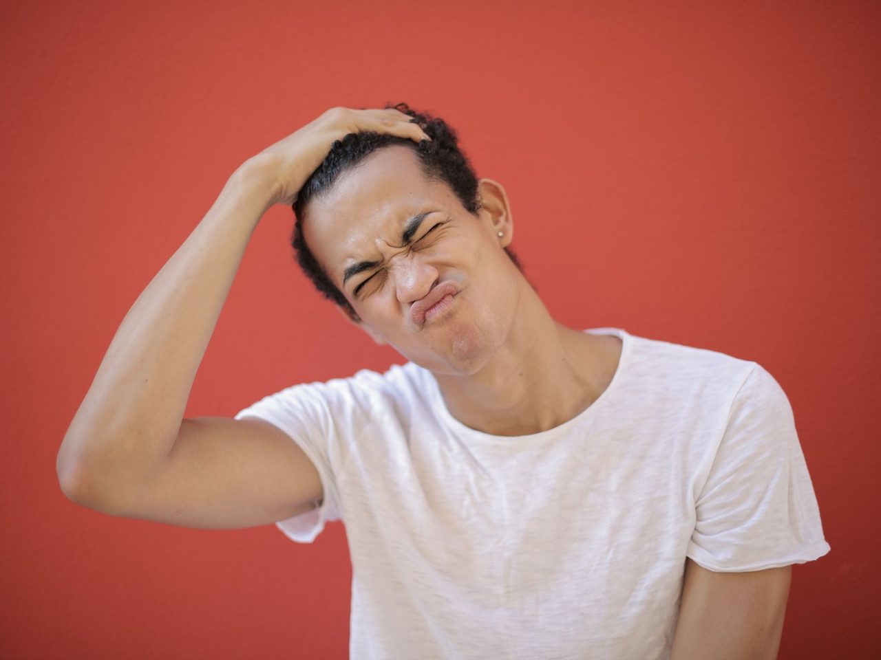 A young man grimacing and scratching his head against a red background