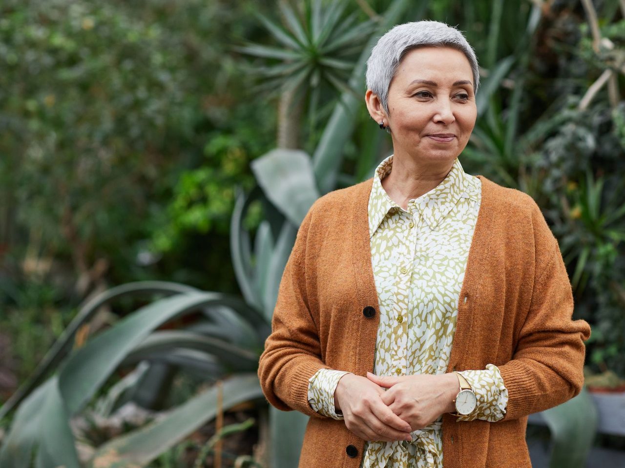 A smiling older woman with short gray hair in a brown cardigan among green plants
