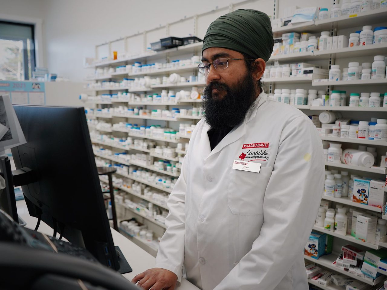 A pharmacist in a white coat and turban working at a computer in a pharmacy