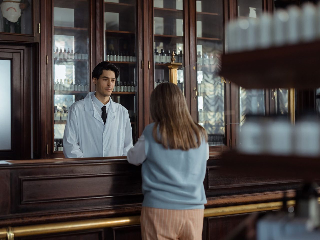 A pharmacist behind an old wooden pharmacy counter talking with a customer