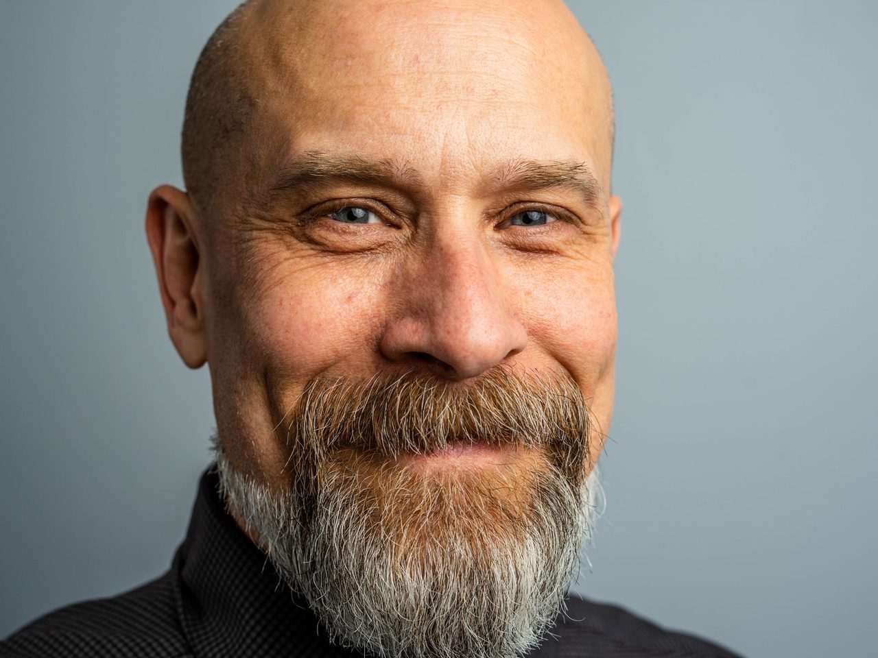 Portrait of a smiling bald man with a full gray beard against a blue-gray background
