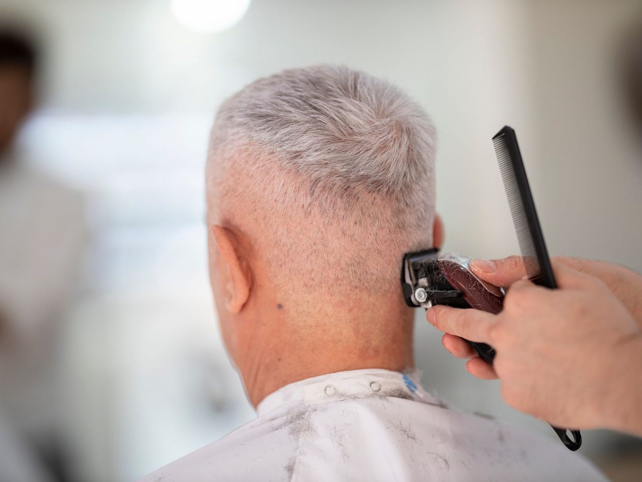 An older man with short gray hair seen from behind while a barber trims it with clippers and a comb