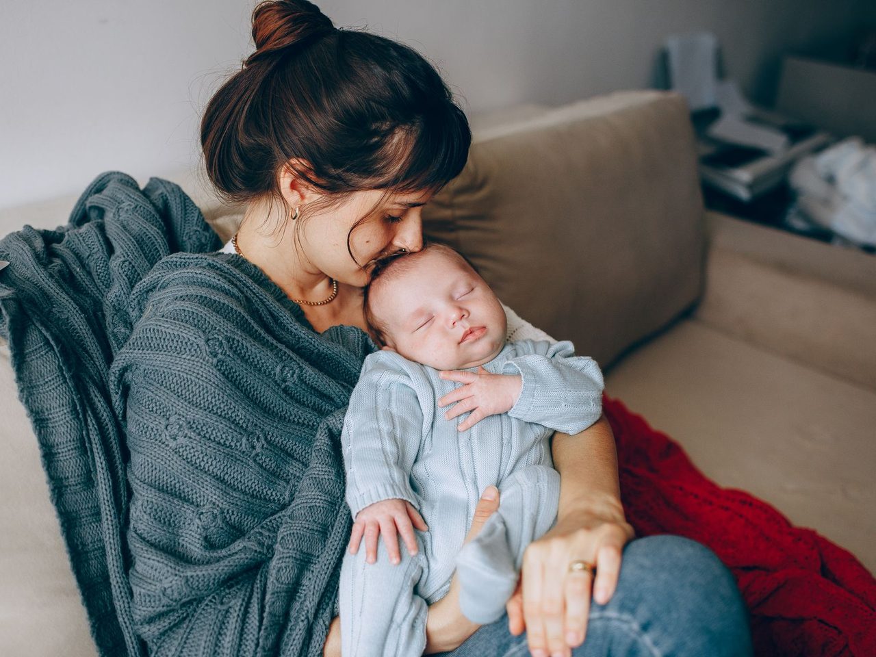 A mother with her hair tied up holding a sleeping baby in a blue sleepsuit on a sofa