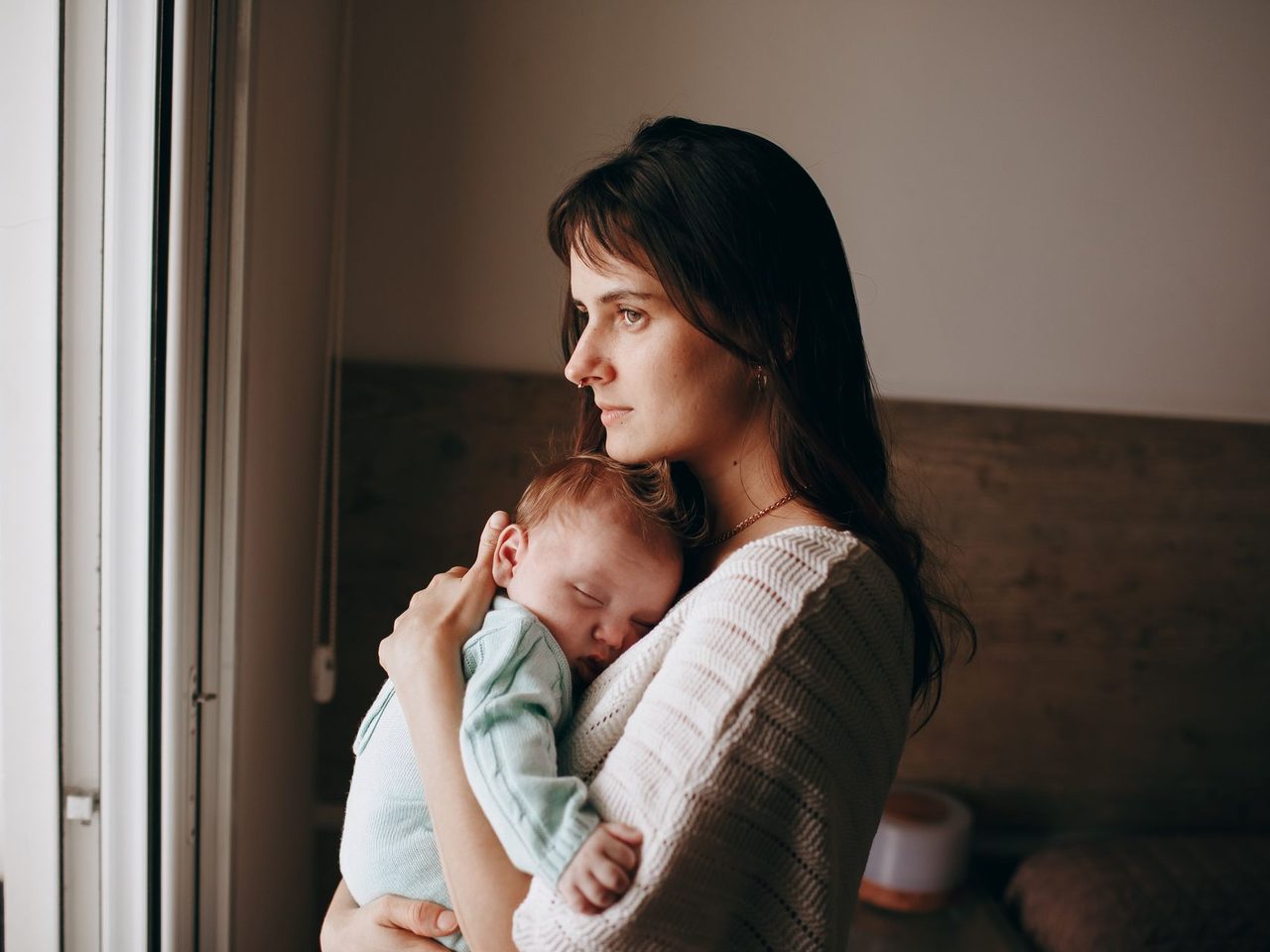 A woman with long dark hair holding a sleeping baby close beside a window