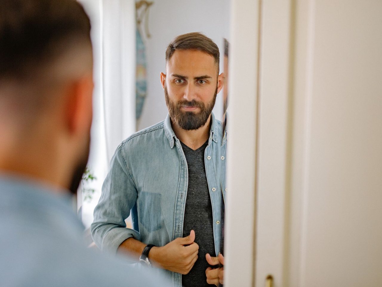 A bearded man adjusting his shirt in front of a mirror at home, seen in the reflection