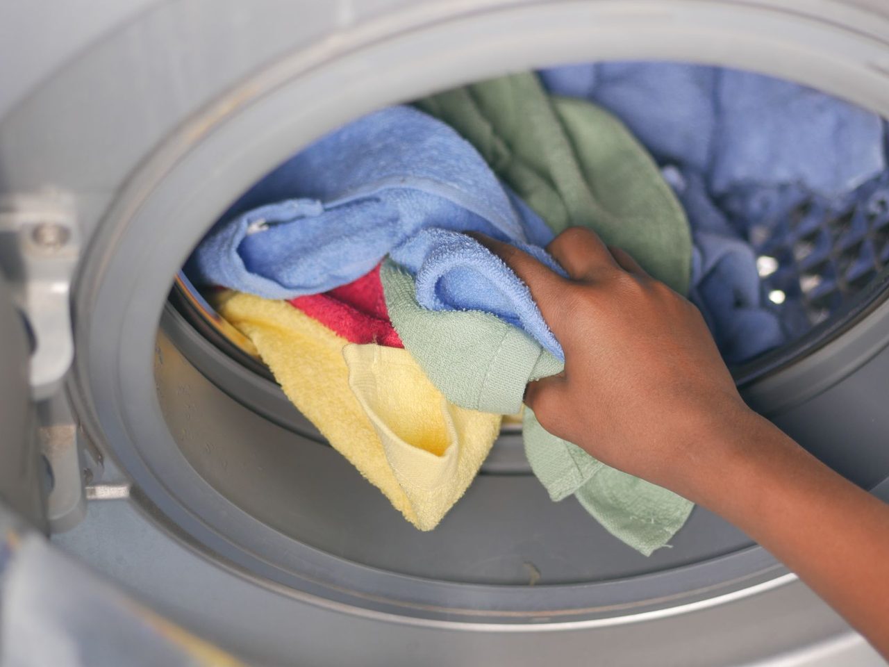 Close-up of a hand placing blue, yellow and green towels into the open drum of a front-loading washing machine.