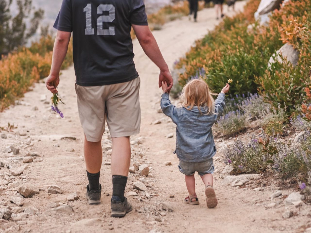 Man in a gray number 12 shirt and shorts holding hands with a small blonde child on a dirt hiking trail, viewed from behind.