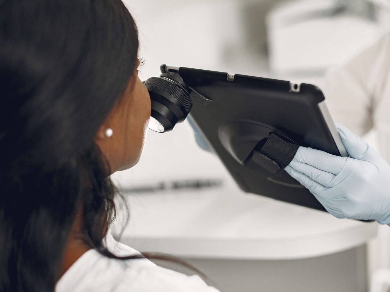 A dermatologist in gloves examining a patient with a digital dermatoscope connected to a tablet