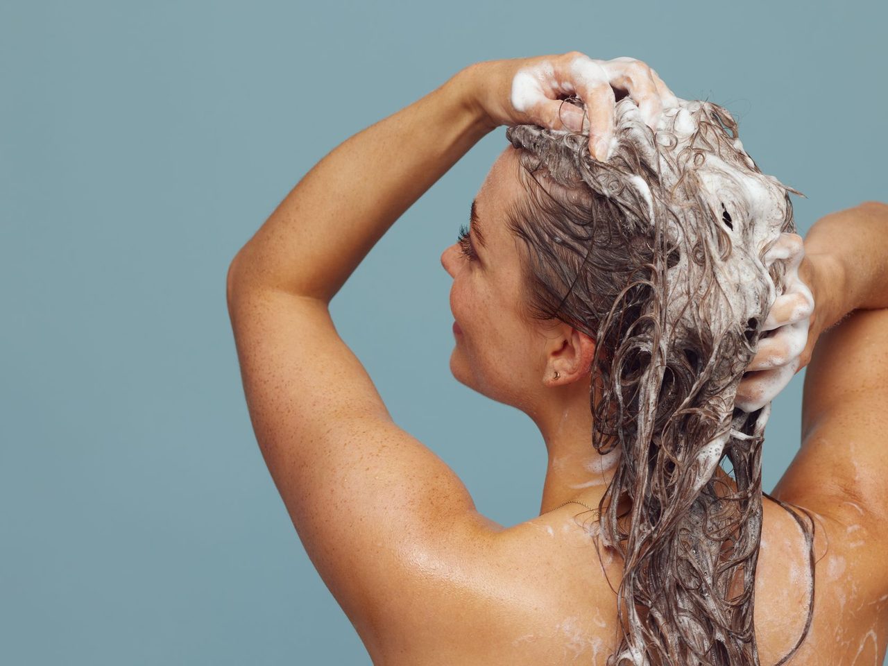 A woman lathering shampoo into her long hair, seen from behind against a blue background