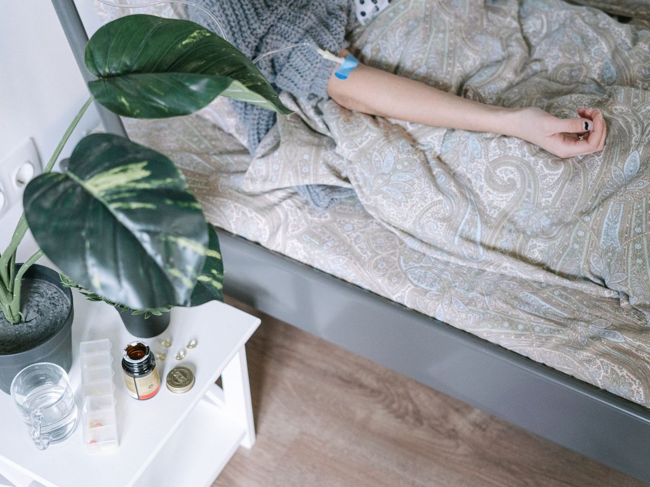 A person lying in bed under a gray blanket with a potted plant and a glass of water on the bedside table