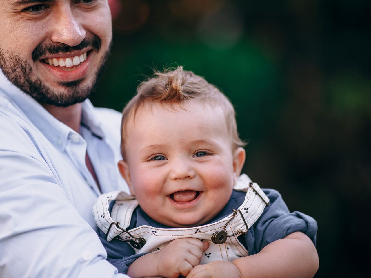 A smiling bearded man holding a laughing baby boy outdoors