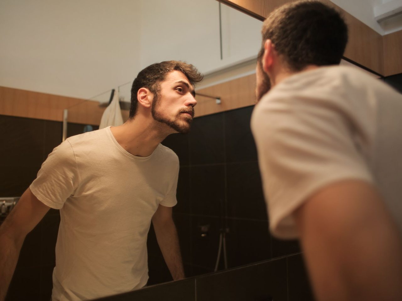 A young man in a T-shirt looking at himself in a bathroom mirror