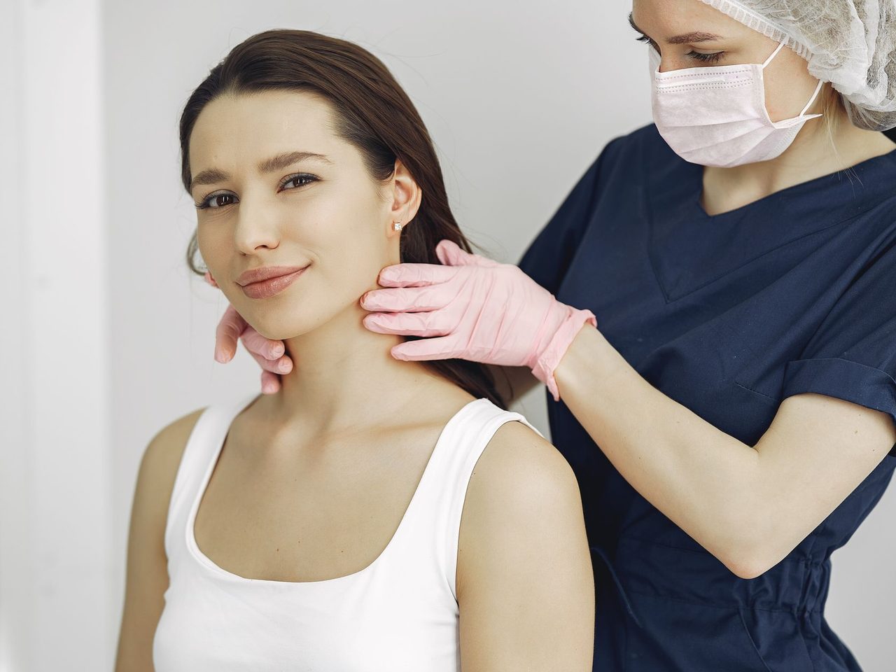 A medical practitioner in scrubs and gloves feeling the front of a smiling woman's neck