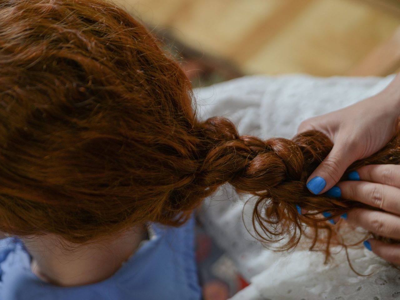 Close-up of a person's hands with blue nail polish braiding long red hair on the back of someone's head.