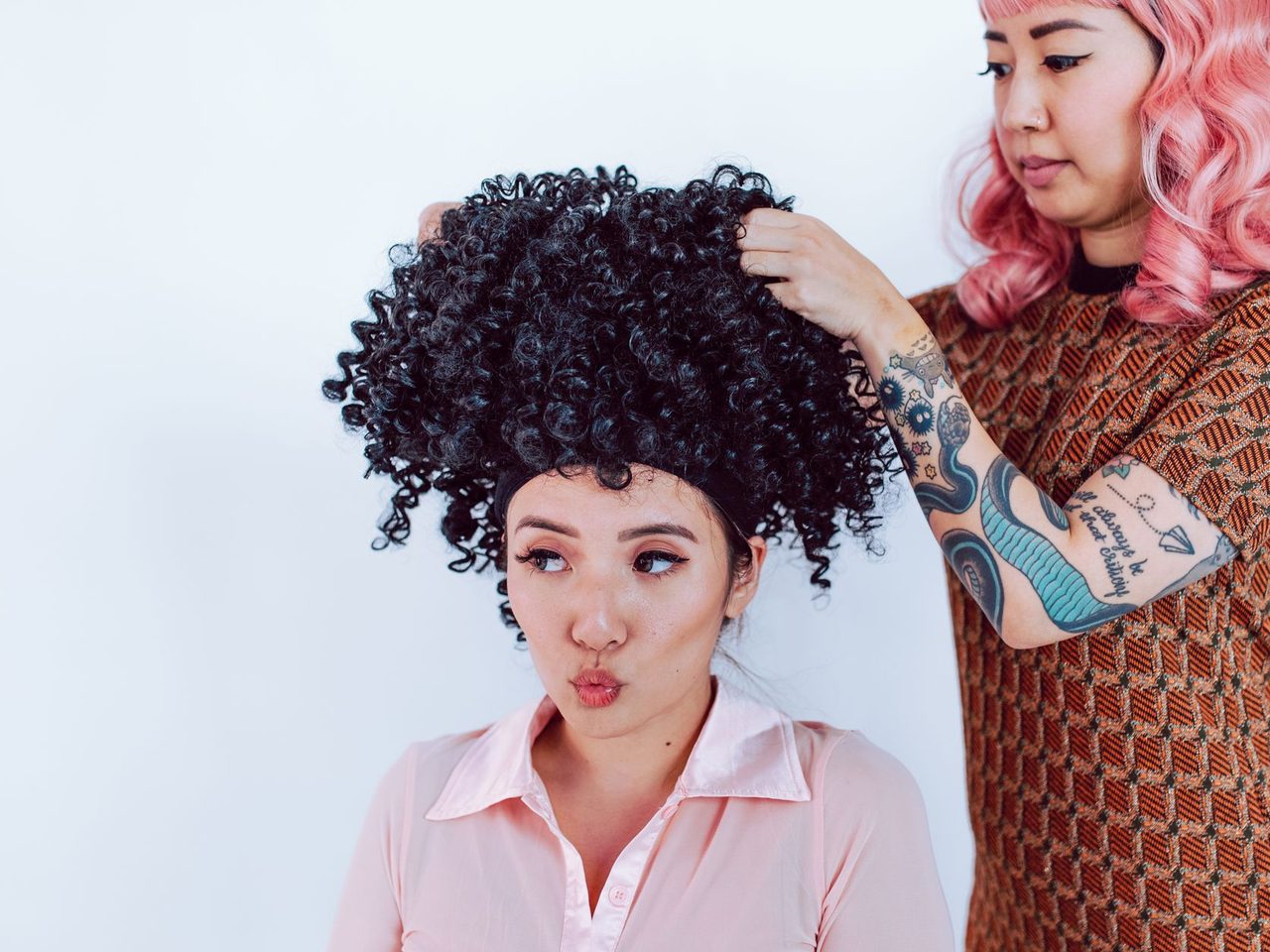 A woman with pink hair and a tattooed arm adjusts a voluminous curly black wig on another woman in a pink shirt.