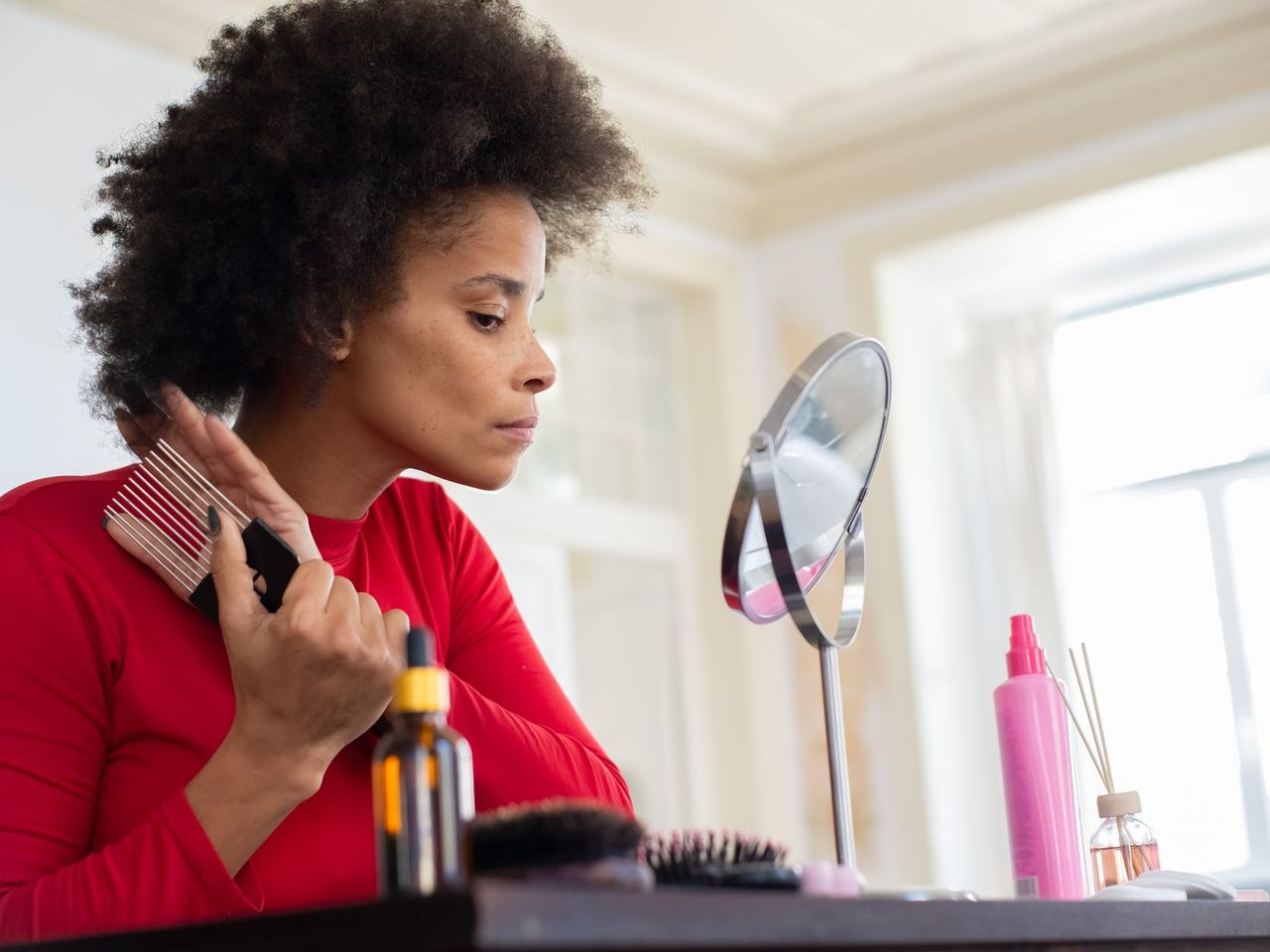 A woman in a red shirt combing her curly hair at a vanity with a round mirror